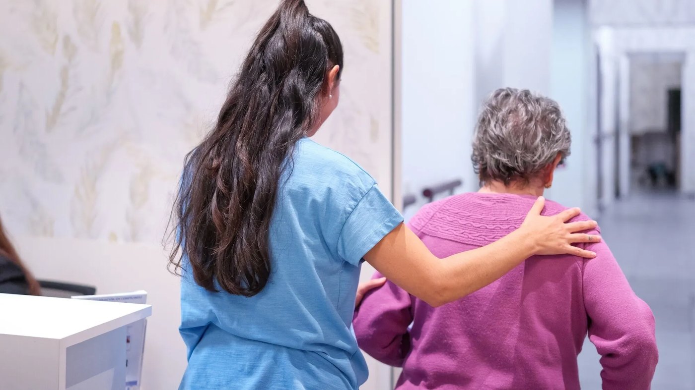 Operatrice che accompagna una signora anziana lungo il corridoio di una struttura sanitaria a Oristano